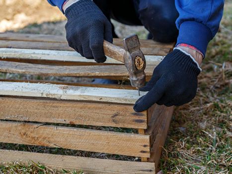 Wooden box made of brown boards on ground, man in black gloves hammers nail into board.