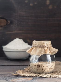 Active rye sourdough starter in glass jar and rye flour on brown wooden background. Starter for sourdough bread. Toned image. Copy space. Vertical