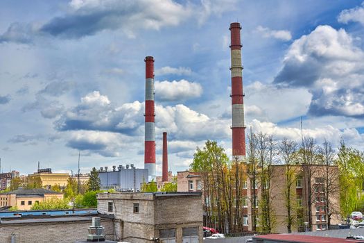 Tall factory chimneys against the backdrop of cloudy sky and residential buildings. Environmental pollution and urban ecology