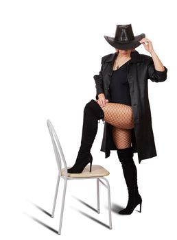 young woman in leather coat, black hat, mesh tights and boots posing standing with her foot on a chair in studio on white background