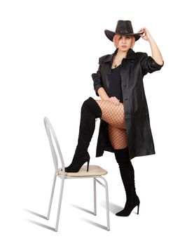 young woman in leather coat, black hat, mesh tights and boots posing standing with her foot on a chair in studio on white background