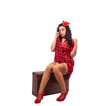 young woman pin-up style in red dress posing sitting on suitcase in studio on white background