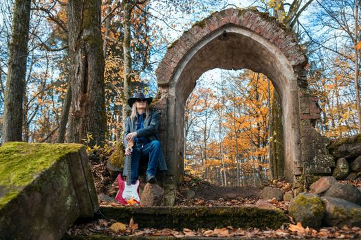 elderly long haired man in a leather coat, hat, sunglasses and jeans standing with an electric guitar in a dark forest near a ruined chapel