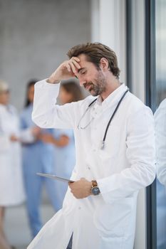 A doctor looking exhaustion while standing near a window and using digital tablet in a hospital hallway during the Covid-19 pandemic.