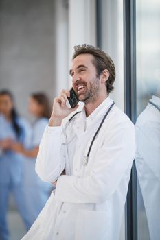 Shoot of a smiling doctor standing and talking on a smartphone while having quick break in a hospital hallway.