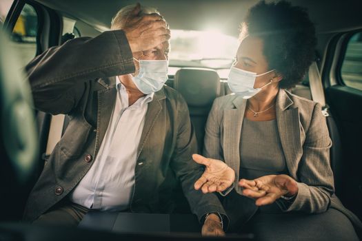 Shot of two worriedly multi-ethnic people with protective mask having a discussion while sitting in the backseat of a car on their business commute during COVID-19 pandemic.