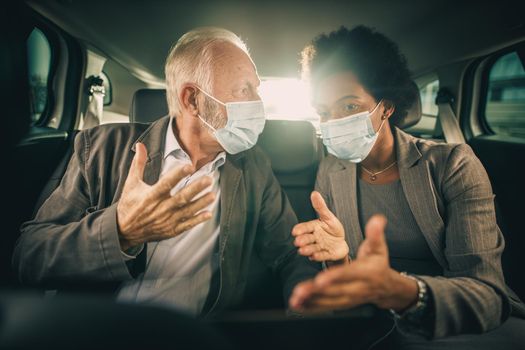 Shot of two multi-ethnic people with protective mask having a discussion while sitting in the backseat of a car on their business commute during COVID-19 pandemic.