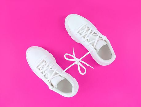 Pair of white sport shoes connected with laces bow on a pink background.