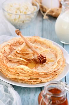 Homemade thin crepe (pancakes) with honey stacked in a stack, on a table with a pot of milk, a bowl of cottage cheese and eggs in a basket. Country style food. Traditional Slavonian, pagan holiday (Maslinitsa)
