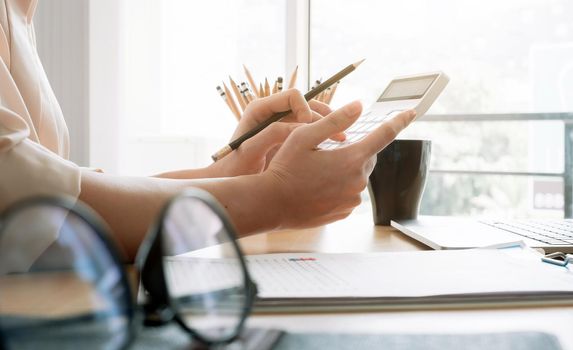 Close up of businesswoman or accountant hand holding pencil working on calculator to calculate financial data report, accountancy document and laptop computer at office, business concept