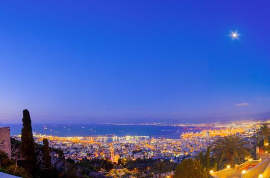 Evening view of the Bahai Shrine and gardens, with the downtown, the port, and the moon, in Haifa, Northern Israel