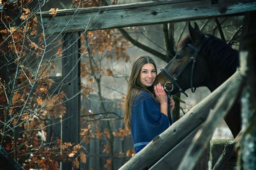 A beautiful girl in a blue stole stands next to a horse on the background of wooden buildings