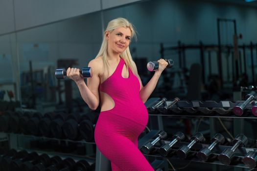 Pregnant woman lifting up dumbbells training biceps muscle at the gym standing near mirror Pregnancy, healthy lifestyle, sport and fitness concept Caucasian female athlete workout training