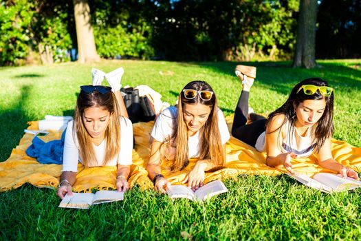 Three young women students spending time lying on meadow in city park at sunset or dawn reading book to learn university school lesson. Girl relaxing in city park enjoying preferred romance tale