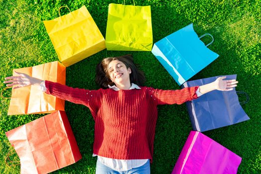 View from top of smiling girl lying on green meadow with open arms among iris colored shopping bags happy for newly purchases. Shopaholic young woman happiness buying. Modern times consumerism concept