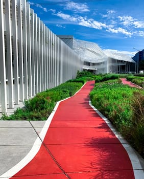 Empty red walkway for running in the garden near shopping mall. Running track in the park with green ornamental plant on sunny day. Public park for exercise and relaxation beside modern building.
