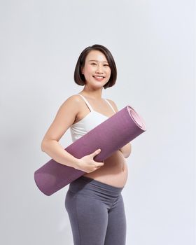 Joyful pregnant woman holding yoga mat smiling to camera posing over white background. Pregnant lifestyle and fitness.