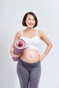 Young attractive pregnant female touching her belly while holding yoga mat in hands, expectant mother doing physical exercises, posing isolated over white background.
