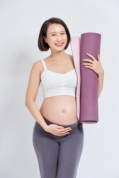 Joyful pregnant woman holding yoga mat smiling to camera posing over white background. Pregnant lifestyle and fitness.