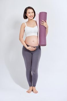 Young attractive pregnant female touching her belly while holding yoga mat in hands, expectant mother doing physical exercises, posing isolated over white background.