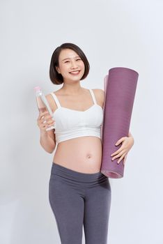 Happy young active pregnant female with bottle of water and rolled mat looking at you with smile while standing over white background