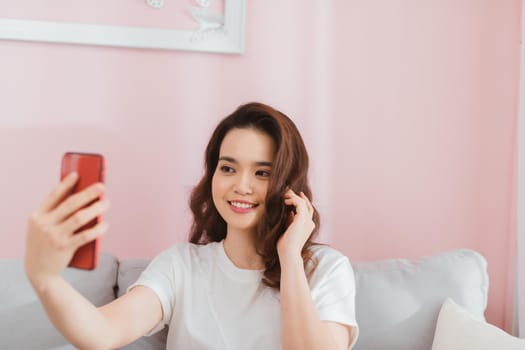 Attractive smiling young asian woman wearing casual clothes sitting on a couch, taking a selfie