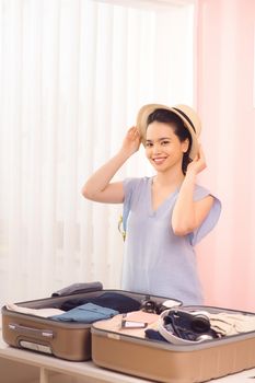 Beautiful girl in stylish clothes and hat is posing at camera and smiling while packing a travel bag at home