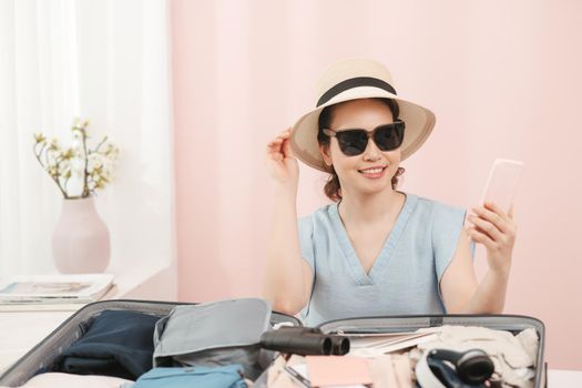 Young woman choosing hat sitting with clothes and suitcase on the bed. Prepairing for the summer vacations