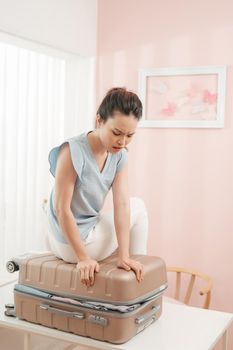 Sad young woman sitting on stuffed suitcase, smiling. Girl packing bag for travel, closing hardly overfilled luggage bag.