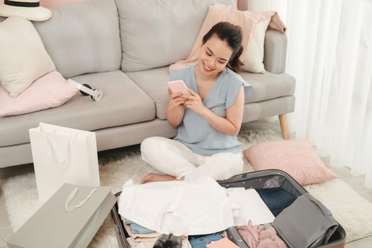 Woman hands using smartphone write checklist to prepare accessories for new journey, packing clothes in suitcase bag