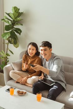 Couple watching tv sitting on a sofa in the living room at home. Man is zapping and the woman is holding a tablet and looking at you