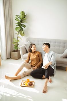 Relaxing at home. Happy young man and woman drinking hot beverage with sweet pastry. They are sitting at table on floor near couch and smiling