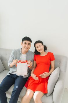 Young husband and a pregnant wife show their baby t-shirt when sitting on sofa.