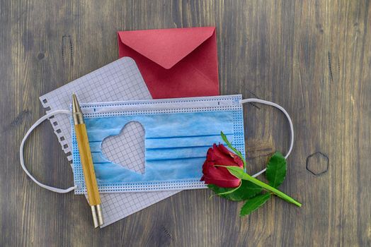 Face mask with cut out heart shape alongside a wire bound notepad and pen and red envelope on wooden background with free copy space. Social distancing and contact-less greetings