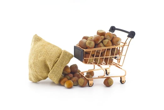 Small hessian bag and shopping cart filled with whole ripe hazelnuts spilling onto a white background
