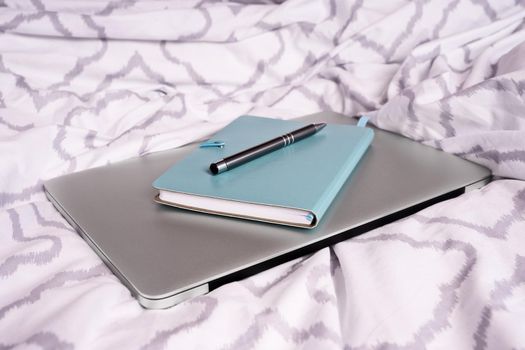 Notepad and pen on top of a laptop computer on bed with modern bright bed sheets. Mobile working concept. Freelance, distance learning or work from home concept.