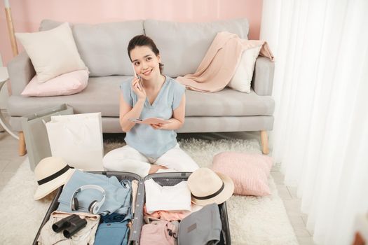 young woman talking with friend on mobile phone holding passport packing travel suitcase organizing clothes and things in summer holiday trip