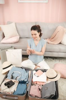 Smiling elegant woman using mobile travel agent app on smartphone near open travel suitcase.