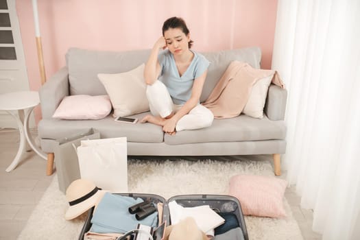 Young woman having trouble packing suitcase bag, sitting on sofa, hand on face concerned, not ready