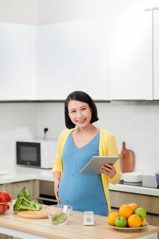 smiling young pregnant woman using digital tablet while cooking in kitchen