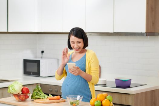 Happy pregnant woman calling on smartphone and having breakfast at home kitchen