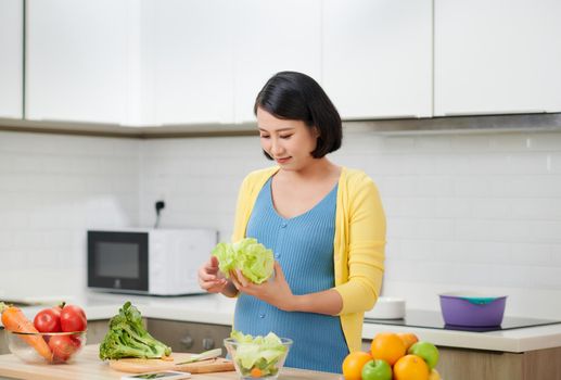 Happy pregnant woman cooking at home, doing fresh green salad, eating many different vegetables during pregnancy, healthy pregnancy concept