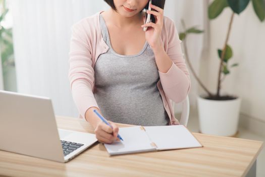 Young cheerful pregnant woman talk on phone. Also working remote using laptop