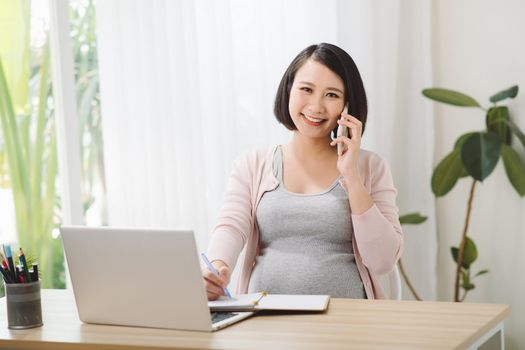 Young cheerful pregnant woman talk on phone. Also working remote using laptop