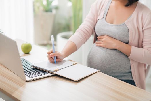 Beautiful pregnant business woman is writing in her notebook and smiling while standing near the working place at home