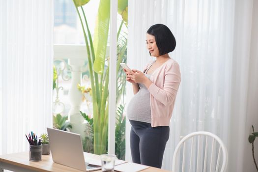 Young pregnant woman standing at window and reading text message on smartphone