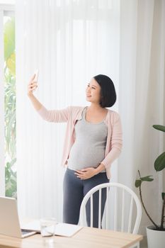 A young pregnant woman standing next to the window taking a selfie