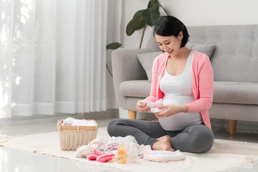 beautiful pregnant asian woman using pen writing list on notebook packing and preparing baby clothes in basket