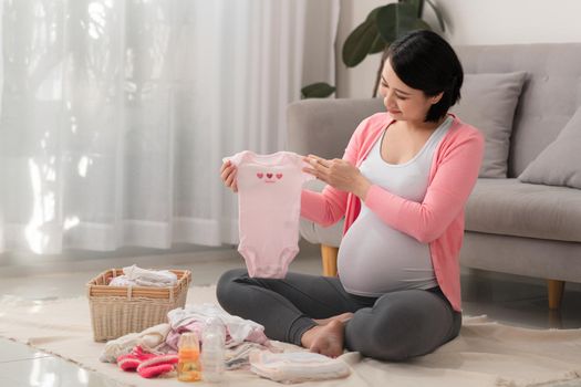 Young pregnant woman holds baby clothes on white background sitting at home on the floor