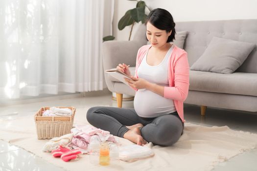 Cheerful pregnant preparing baby clothes and making to do list, getting ready for baby delivering